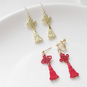 May include: A set of three pairs of earrings featuring a gold and red tassel with a knot design. The earrings are available in both clip-on and post styles.