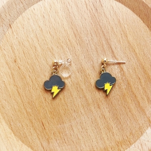 Puede incluir: Pendientes con un diseño de nube gris oscuro y rayo amarillo. Un pendiente tiene un poste, el otro un clip. Los pendientes se exhiben sobre una superficie de madera clara.