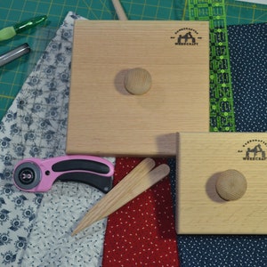 May include: Two light-colored wooden blocks with a knob, a pink rotary cutter, and other quilting tools are arranged on a green cutting mat. The blocks have the text "Woodcraft" and a logo. Fabric pieces in blue, white, and red are also present.