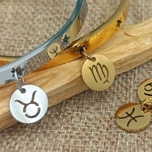 Peut inclure: Deux bracelets en or et en argent avec des breloques du zodiaque. Les breloques sont pour le Taureau, la Vierge et les Poissons. Les bracelets sont sur une surface en bois.