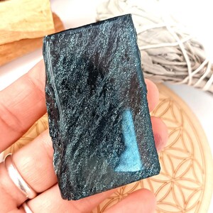 Peut inclure: Un cristal rectangulaire bleu foncé avec une surface texturée. Le cristal est tenu dans une main, avec un dessous de verre en bois et un paquet de sauge en arrière-plan. Une statue de Bouddha en bronze est également visible.