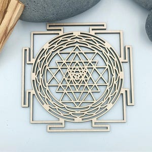 Puede incluir: Se muestra un Sri Yantra de madera, un símbolo geométrico sagrado. El diseño presenta triángulos entrelazados que forman un patrón central, rodeado por un marco cuadrado. La imagen también incluye piedras grises lisas y varios trozos de madera de color marrón claro.