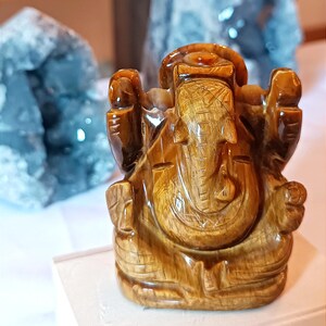 May include: A detailed, carved wooden statue of Ganesha, the Hindu god of wisdom, with a prominent elephant head and multiple arms. The statue is a warm brown color and sits in front of two large, light blue crystal formations.