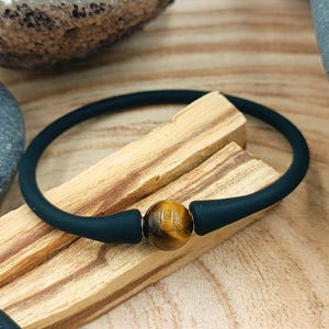 Peut inclure: Un bracelet en silicone noir avec une perle ronde en œil de tigre brun doré. Le bracelet est posé sur deux morceaux de bois clair. Le fond est une surface en bois brun clair.