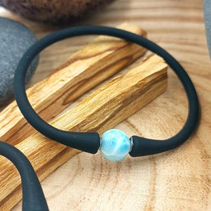 Peut inclure: Un bracelet en silicone noir avec une perle de pierre précieuse bleu clair et blanche. Le bracelet est présenté sur une surface en bois avec deux morceaux de bois brun clair. La pierre précieuse est ronde et a un aspect marbré.