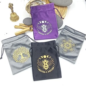Può includere: Quattro sacchetti in velluto con coulisse in viola, nero e grigio. Ogni sacchetto presenta una grafica dorata o argentata diversa: un'ape, un albero della vita e un Cubo di Metatron. Una piccola campana di ottone e bastoncini di incenso sono sullo sfondo.