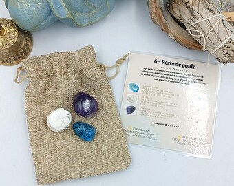 May include: A small burlap pouch containing three tumbled stones: a purple amethyst, a white howlite, and a blue apatite. A card with the text "6- Perte de poids" is visible in the background.