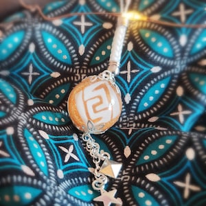 Peut inclure: Un collier de perles dorées et blanches avec un pendentif représentant un symbole blanc sur fond doré. Le pendentif est attaché à une chaîne argentée avec une étoile et un pendentif en forme de pyramide.