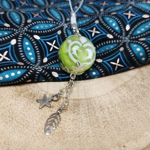 Op de afbeelding: Een groene glazen bedel met een wit hartontwerp en zilveren accenten hangt aan een zilveren ketting met ster- en bladbedels. De bedel ligt op een houten oppervlak met een blauw-zwarte gepatenteerde stof op de achtergrond.