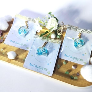 Op de afbeelding: Drie Final Fantasy XIV hars sleutelhangers met goudkleurige kettingen worden gepresenteerd op een houten dienblad. Elke sleutelhanger heeft een blauw en wit ontwerp. De sleutelhangers zijn verpakt op kaarten met het logo van de game en decoratieve elementen.