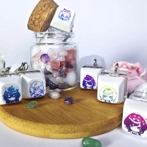 Może przedstawiać: Pięć białych breloków do klawiszy z kolorowymi postaciami w stylu anime nadrukowanymi na nich. Breloki wiszą na srebrnym łańcuszku i są prezentowane na drewnianej powierzchni.