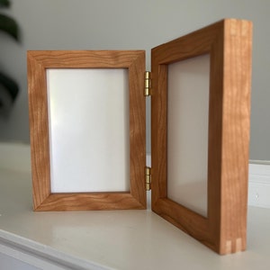 May include: A wooden, double-hinged picture frame with a natural cherry finish. The frame is open, revealing two white spaces for photos. The hinges are brass-colored. The frame is displayed on a white surface.