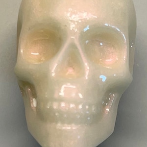 Puede incluir: Figura de calavera nacarada pálida con rasgos faciales detallados. La calavera tiene cuencas oculares visibles, una cavidad nasal y una mandíbula definida. La superficie refleja la luz, creando reflejos y sombras.