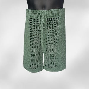 May include: Sage green crochet shorts with a drawstring waist. The shorts feature a lattice design on the front and back. The shorts are made of a soft, breathable material, perfect for warm weather. The shorts are a casual style.