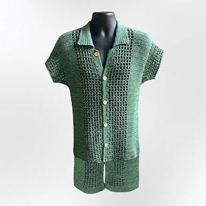 May include: A forest green crochet two-piece set consisting of a short-sleeved button-down shirt with a collar and matching shorts. The garment features an open-weave grid pattern combined with solid crochet panels and light-colored round buttons.
