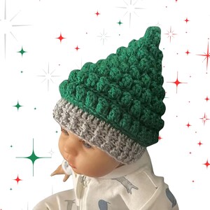 May include: A green and grey crocheted Christmas tree hat. The hat has a textured, bobble stitch pattern in green, resembling a Christmas tree. The brim is grey. The hat is likely for a baby or small child, perfect for the festive season.