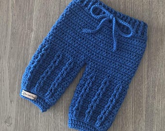 May include: A pair of royal blue crochet pants featuring a ribbed waistband with a matching drawstring tie. The legs display a vertical cable-knit texture pattern. A small rectangular fabric tag is attached to the lower left leg cuff.
