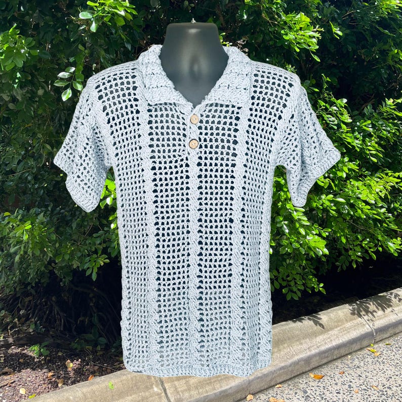 May include: A light blue crochet polo shirt with short sleeves and a collared neckline. The shirt features a button-up front with two wooden buttons. The open weave design is visible throughout the shirt, creating a breathable and textured look.