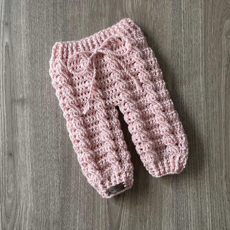 May include: Light pink crochet pants featuring a textured cable knit pattern throughout the legs. The garment includes an elasticized waistband with a matching pink drawstring tie and a small rectangular metallic tag attached at the bottom hem.
