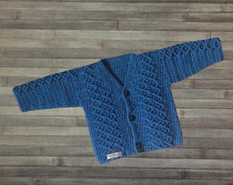 May include: A blue long-sleeved cardigan featuring a textured diamond knit pattern, a V-neckline, and three dark buttons down the front. A small rectangular label is attached to the bottom left corner of the garment.