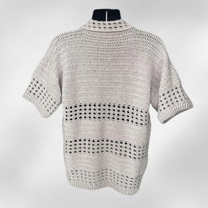 May include: A light beige crochet shirt with short sleeves and a collared neckline. The shirt features a horizontal openwork design across the chest. The crochet work is visible and detailed, creating a textured appearance. The shirt is displayed on a black mannequin.