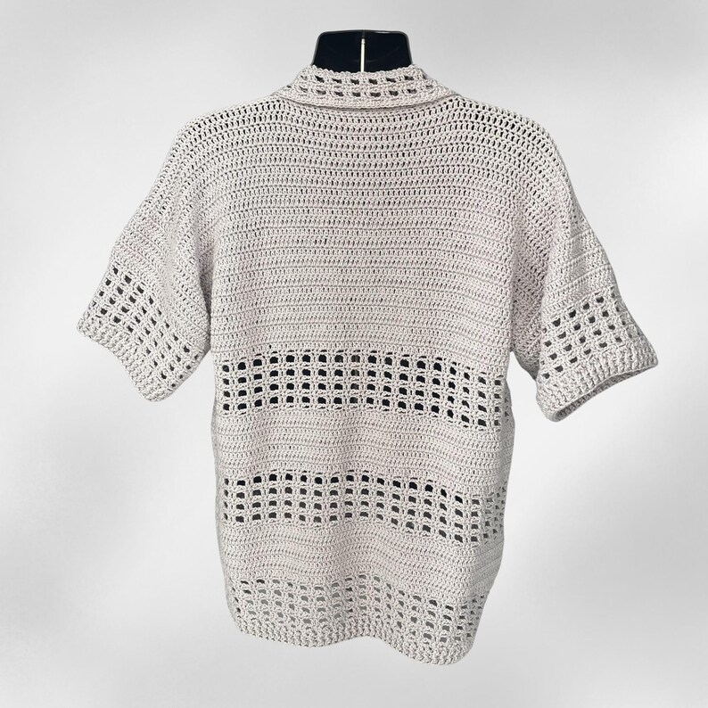 May include: A light beige crochet shirt with short sleeves and a collared neckline. The shirt features a horizontal openwork design across the chest. The crochet work is visible and detailed, creating a textured appearance. The shirt is displayed on a black mannequin.