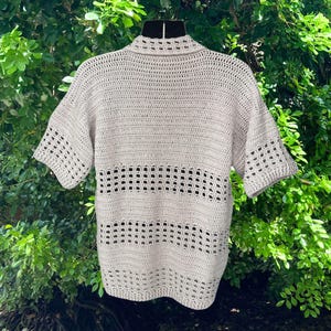 May include: A light beige, short-sleeved crochet top. The top features a collared neckline and openwork detailing across the chest and on the sleeves. The garment is displayed on a black mannequin against a backdrop of green foliage.