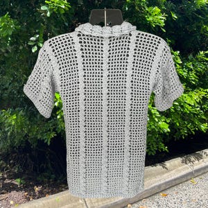 May include: A light gray, short-sleeved crochet shirt. The shirt features a collared neckline and a textured, open weave design. The crochet pattern includes vertical lines and a scalloped edge on the collar. The shirt is displayed on a black hanger.