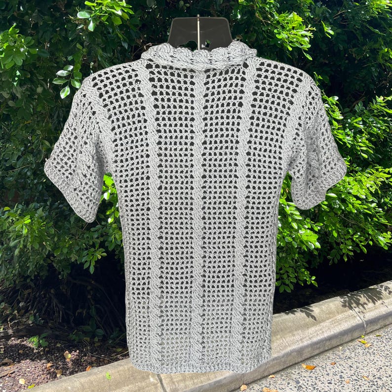 May include: A light gray, short-sleeved crochet shirt. The shirt features a collared neckline and a textured, open weave design. The crochet pattern includes vertical lines and a scalloped edge on the collar. The shirt is displayed on a black hanger.