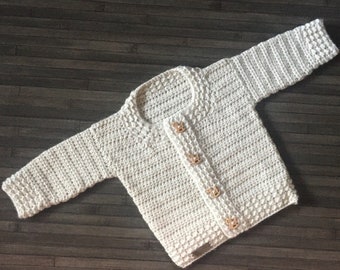 Puede incluir: Una chaqueta de ganchillo color crema de manga larga con cuello redondo y cuatro botones de madera marrón claro en la parte delantera. La prenda está confeccionada con un patrón de punto texturizado e incluye una pequeña etiqueta rectangular en el dobladillo.
