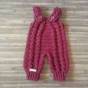 May include: A dark magenta crochet romper featuring a vertical cable knit pattern, two shoulder straps secured with matching buttons, and ribbed cuffs at the leg openings. A small rectangular white label is attached to the lower left leg.