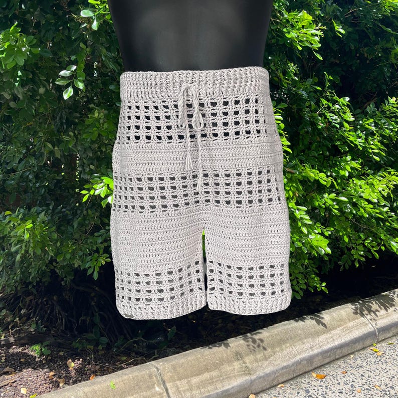 May include: Light gray crochet shorts with a drawstring waist. The shorts have a lattice design and are displayed on a mannequin. The shorts are made of a soft, textured yarn and are suitable for warm weather.