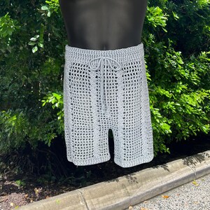 May include: Light gray crochet shorts with a drawstring waist. The shorts have a textured, open weave design and are displayed on a mannequin. The shorts are knee-length and appear to be handmade.