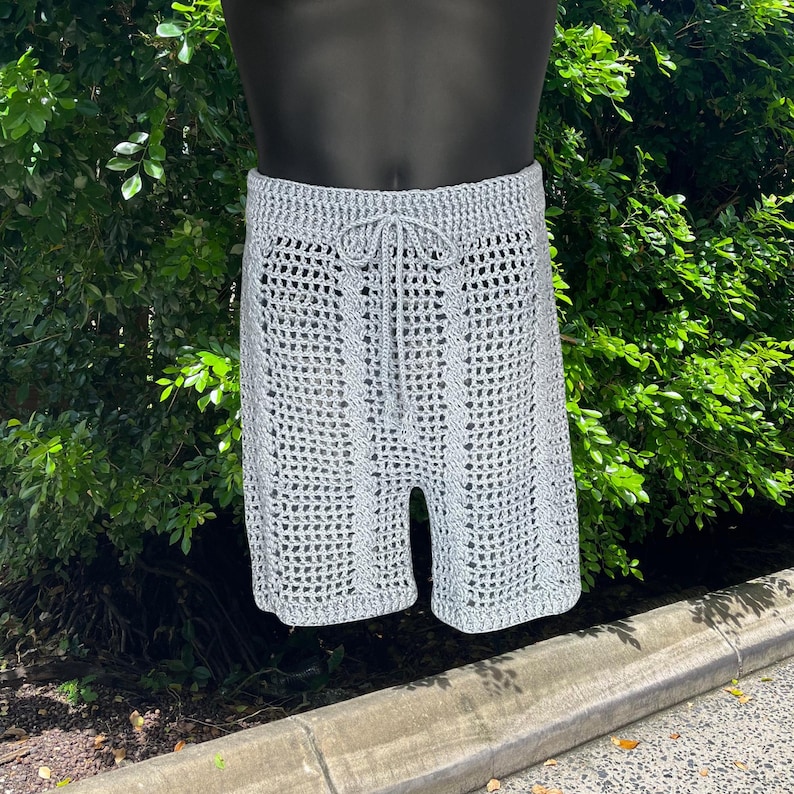 May include: Light gray crochet shorts with a drawstring waist. The shorts have a textured, open weave design and are displayed on a mannequin. The shorts are knee-length and appear to be handmade.