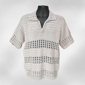May include: A light beige, short-sleeved crochet shirt with a V-neck collar. The shirt features a textured, open-weave design with horizontal bands of larger open spaces. The shirt is displayed on a black mannequin.