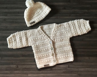May include: A cream-coloured crocheted long-sleeve cardigan with a V-neckline, four front buttons, and a matching beanie hat topped with a pom-pom. Both items feature a textured stitch pattern and a small rectangular tag on the hem.