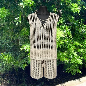 May include: A beige crochet two-piece set consisting of a sleeveless top and shorts. The top features a V-neckline with crisscross lace-up detailing and small dark brown wooden beads. The entire set is crafted in an open-knit mesh pattern.