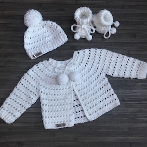 Könnte beinhalten: Ein weißes Häkelset für Babys, bestehend aus einer langärmeligen Strickjacke mit zwei Bommeln am Ausschnitt, einer passenden Mütze mit einem Bommel oben und einem Paar Babyschuhen mit Bindebändern. Jedes Teil hat ein kleines rechteckiges Metallschild.