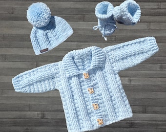 Peut inclure: Ensemble de crochet pour bébé bleu clair, comprenant un chapeau avec un pompon, un cardigan avec des boutons en bois et des chaussons. L'ensemble est présenté sur une surface en bois. Le chapeau a une petite étiquette marron.