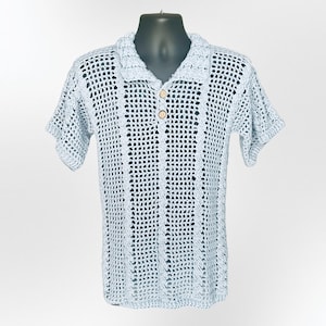May include: A light blue crochet polo shirt with short sleeves and a collared neckline. The shirt features a button-down front with two wooden buttons. The open weave design is visible throughout the shirt, creating a textured appearance.