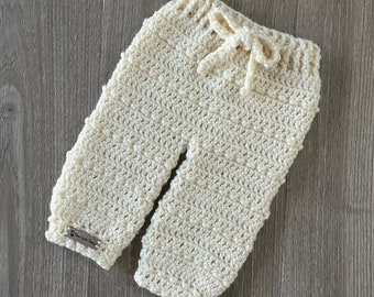 Puede incluir: Pantalones de ganchillo color blanco roto con cintura acanalada y cordón funcional. La prenda presenta un patrón de puntos texturizado y una pequeña etiqueta rectangular marrón en el puño de la pierna izquierda que dice "Lana's Crochet Designs."