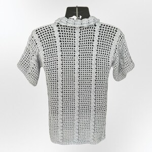 May include: A light gray, short-sleeved crochet shirt. The shirt features a textured, open weave design with a ruffled collar. The garment is displayed on a black mannequin, showcasing the back of the shirt against a white background.