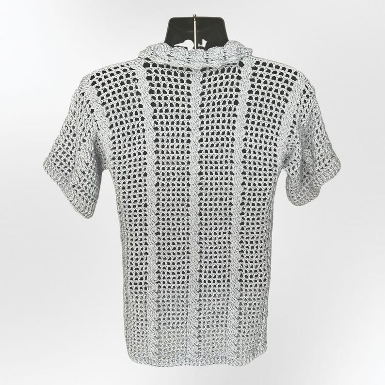 May include: A light gray, short-sleeved crochet shirt. The shirt features a textured, open weave design with a ruffled collar. The garment is displayed on a black mannequin, showcasing the back of the shirt against a white background.