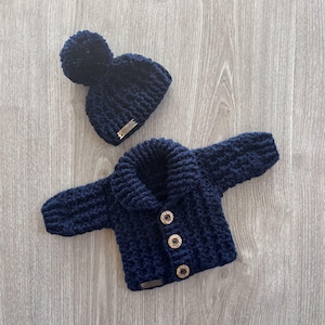 Può includere: Un cardigan all'uncinetto blu navy scuro con collo a scialle e tre bottoni in legno marrone chiaro, abbinato a un berretto all'uncinetto blu navy coordinato con un grande pompon sulla sommità e una piccola etichetta metallica rettangolare.