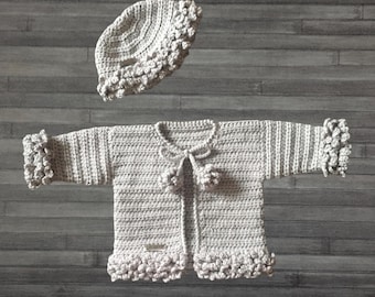 May include: A light grey crochet cardigan and matching hat set. The cardigan features long sleeves with ruffled cuffs, a ruffled hem, and a front tie closure with two pom-poms. The hat has a matching ruffled brim and textured crochet pattern.