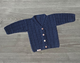 May include: A dark navy blue long-sleeve cardigan featuring a textured crochet pattern, a V-neckline, and four light-colored circular buttons down the front. A small rectangular label is attached to the bottom left corner of the garment.