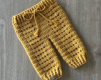 May include: Mustard yellow crochet pants featuring a textured stitch pattern, a ribbed waistband with a matching drawstring tie, and a small rectangular metal brand tag attached to the bottom hem of the left pant leg.