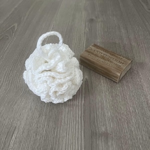 May include: White crocheted bath pouf with a loop handle, next to a bar of sandalwood soap with the text "BOTANICALS DAY SANDALWOOD NATURAL SOAP WITH PURE PLANT OIL".
