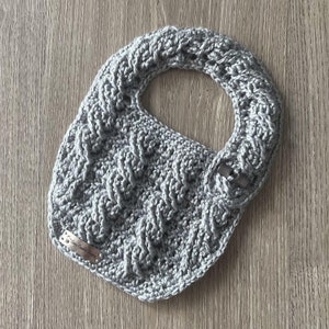 Può includere: Un bavaglino per neonato in crochet grigio con un motivo testurizzato. Il bavaglino ha un'etichetta piccola con il testo "Handmade with Love".