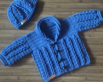 May include: A bright blue crocheted long-sleeve cardigan with a shawl collar and four dark toggle buttons down the front. The set includes a matching blue crocheted beanie hat. Both items feature a textured cable knit pattern throughout.